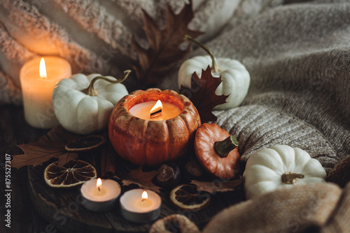 Autumn composition with burning candles, mini munchkin pumpkins, warm wool knitted sweater on the wooden windowsill. Dark colours. Cozy home atmosphere, Thanksgiving decor, fall inspiration. Close up