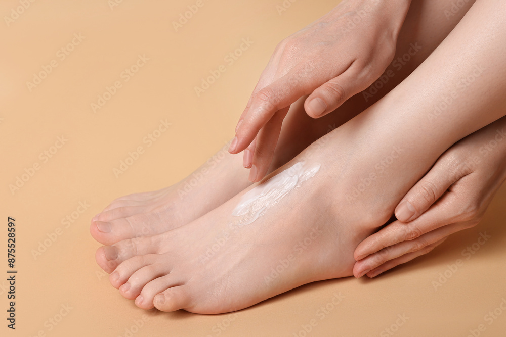 Fototapeta premium Woman applying moisturizing cream onto her feet on beige background, closeup. Body care
