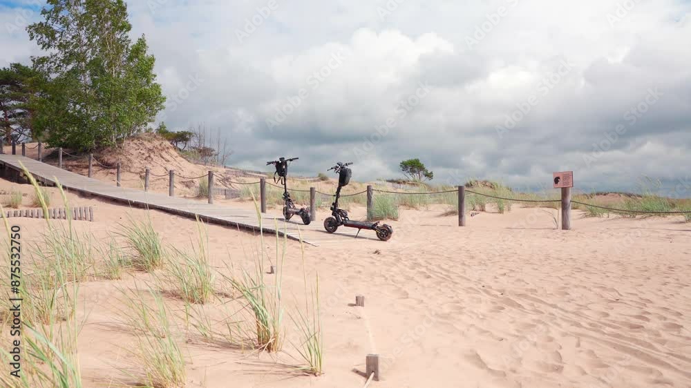 Two electric scooters are parked on a sandy beach with a wooden boardwalk leading to the dunes. Tall grass grows around the boardwalk, and the sky is cloudy. Two electric scooters are parked on a sandy beach with a wooden boardwalk leading to the dunes. Tall grass grows around the boardwalk, and the sky is cloudy.