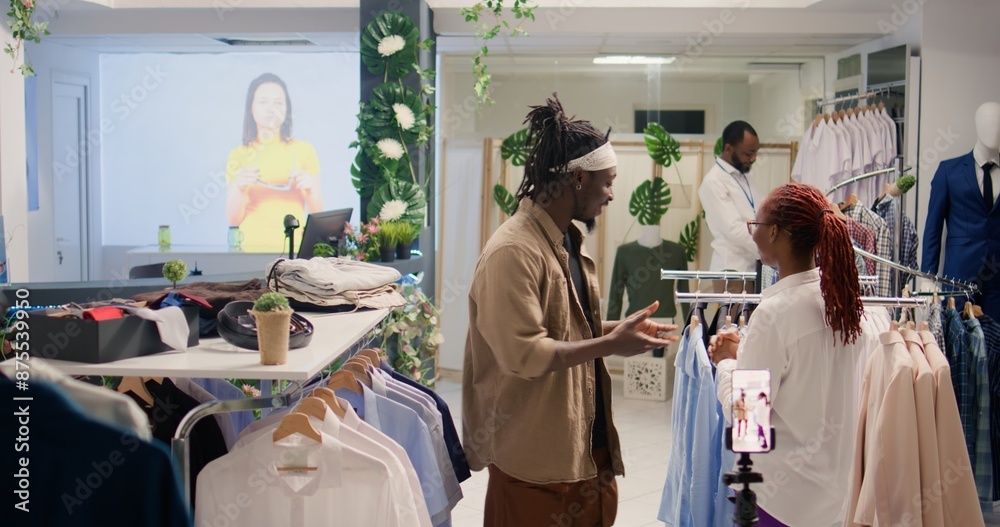 Media star filming social media content in thrift shop with retail assistant, asking her to show followers hidden gems. Influencer in SH clothing store doing shopping haul helped by employee