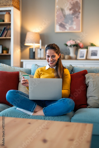 Beautiful smiling woman with laptop and credit card sitting on couch in living room at home.