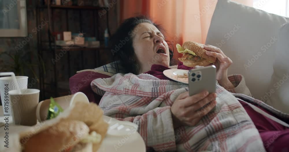 Lazy woman yawning, lying on couch with smartphone, eating fast food burger. Watching bored content. Lazy, careless concept. Negative lifestyle choices. Empty, meaningless, harmful lifestyle