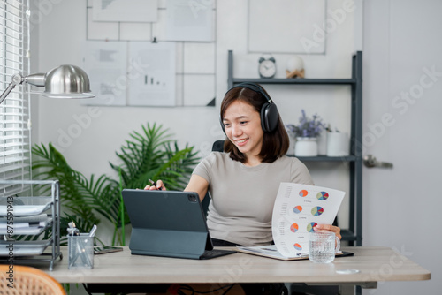 Young woman wearing headphones, working on a tablet, analyzing charts in a modern home office setting with plants and shelves.