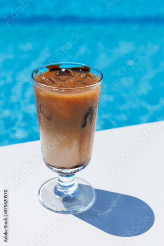 Iced coffee Frappe Fredo against blue clear water of swimming pool, on white table . Summer minimalistic background, holiday or vacation concept. Sun and shadows. Close up, vertical photo