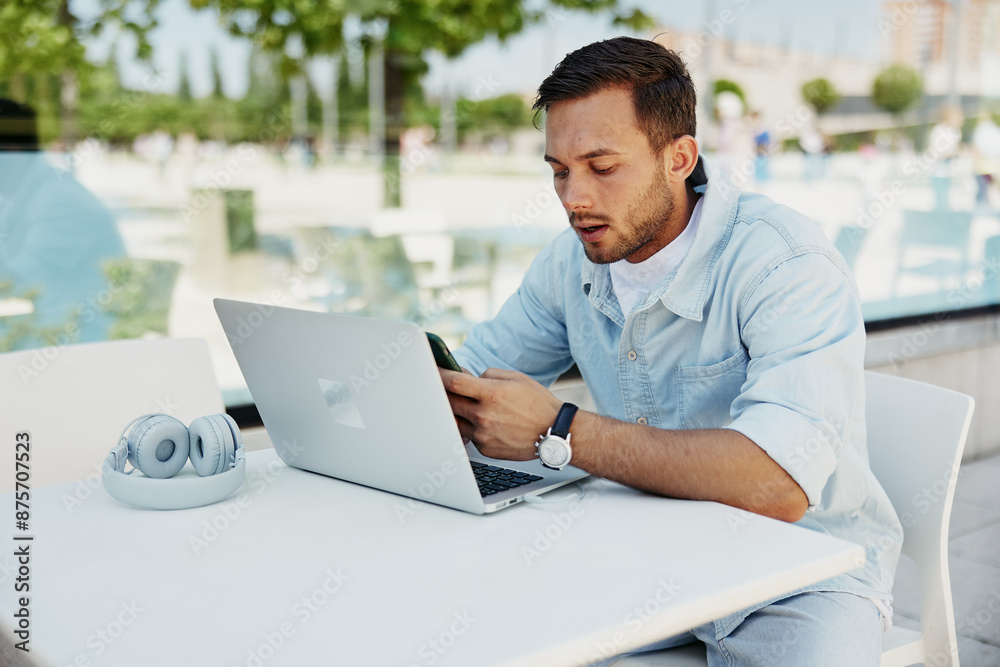 Young man focused on smartphone while working on laptop at outdoor cafe, illustrating multitasking and modern technology Young man focused on smartphone while working on laptop at outdoor cafe, illustrating multitasking and modern technology