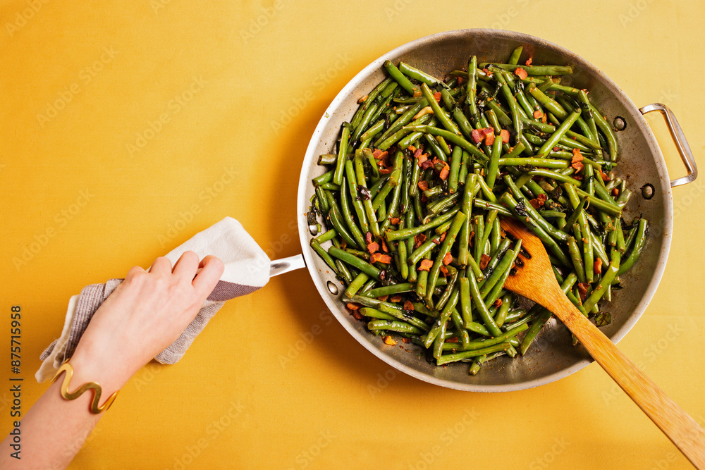 Pan of Green Beans and Bacon on a Yellow Table Cloth with Hand and Spoon Pan of Green Beans and Bacon on a Yellow Table Cloth with Hand and Spoon