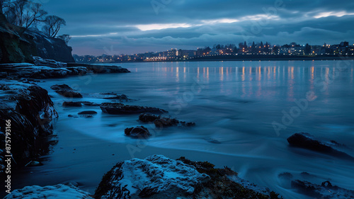 Stony shore and smooth water under a cloudy sky. Seascape with distant city lights reflected in the quiet surface. Peaceful atmosphere of the evening coastline