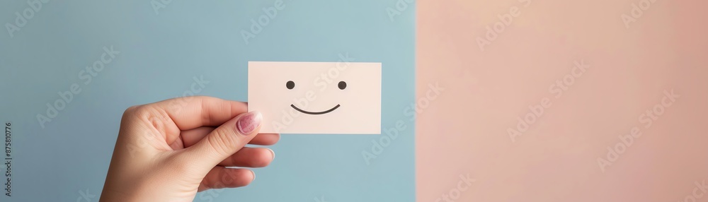 Obraz premium A hand holding a small card with a smiley face in front of a split color background featuring blue and peach tones, symbolizing positivity.
