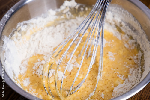 Bowl of batter and whisk on wooden table. Close-up of mixing dough for bakery and cooking. Ingredients for waffles, cakes or pancakes. Dough preparation process