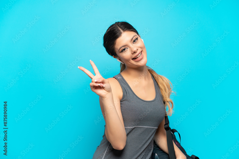 Young sport Russian girl with sport bag isolated on blue background smiling and showing victory sign Young sport Russian girl with sport bag isolated on blue background smiling and showing victory sign