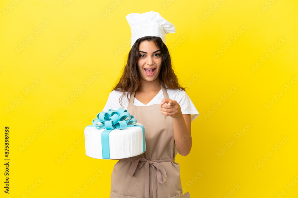 Young caucasian pastry chef woman with a big cake isolated on yellow background surprised and pointing front Young caucasian pastry chef woman with a big cake isolated on yellow background surprised and pointing front