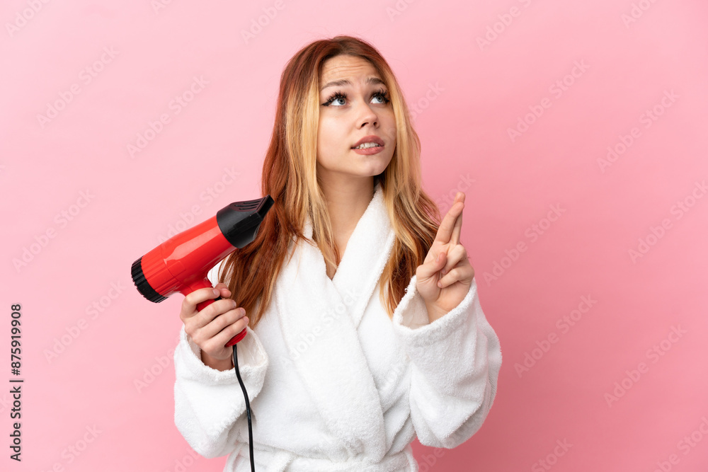 Teenager blonde girl holding a hairdryer over isolated pink background with fingers crossing and wishing the best Teenager blonde girl holding a hairdryer over isolated pink background with fingers crossing and wishing the best