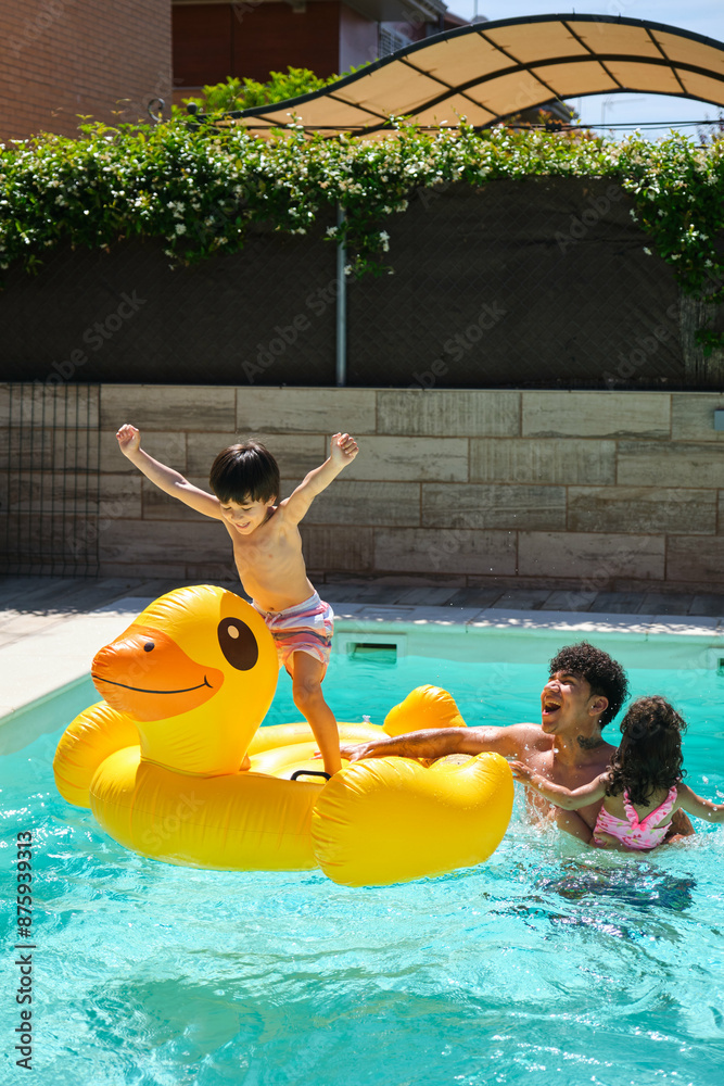 A boy standing on an inflatable duck playing in a pool with his father and sister. The father is holding the duck while the sister is hugging him. The scene is lighthearted and fun
