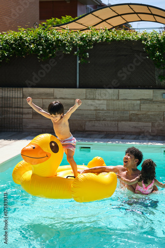 A boy standing on an inflatable duck playing in a pool with his father and sister. The father is holding the duck while the sister is hugging him. The scene is lighthearted and fun