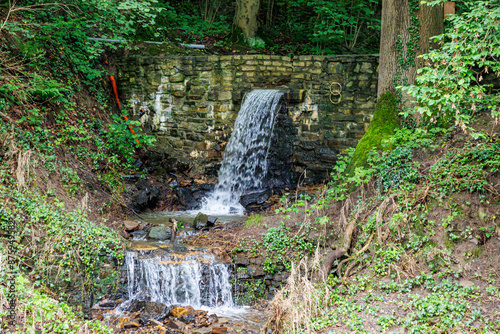 Flowing water coming out of canal between wall, falling into a stream with stones and surrounded by green wild sparse vegetation, cloudy summer day in gardens of Val-Dieu Abbey, in Aubel, Belgium photo