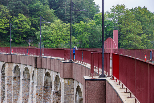 Robertville Dam with red metal fence, rural road for vehicles and pedestrians on top of crest and huge arched wall, green leafy trees on blurred and foggy background, cloudy day in Waimes, Belgium photo