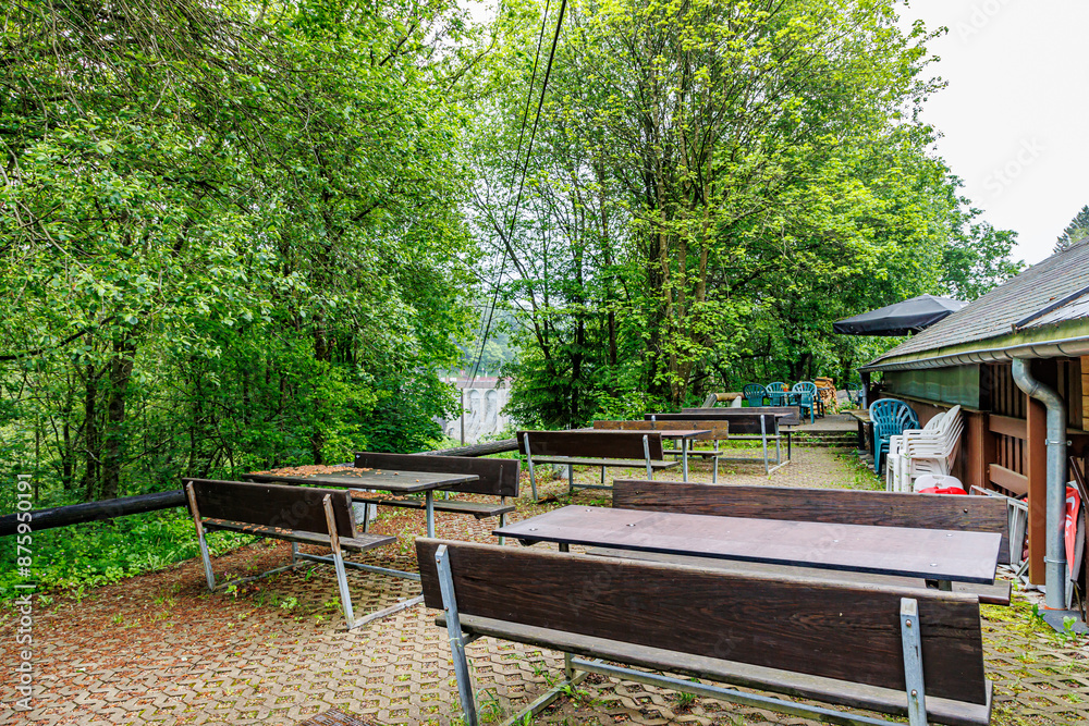 Rustic back terrace of a country restaurant, wooden tables and benches, plastic chairs stacked next to wall, leafy trees with green foliage in background, cloudy summer day in Waimes, Belgium Rustic back terrace of a country restaurant, wooden tables and benches, plastic chairs stacked next to wall, leafy trees with green foliage in background, cloudy summer day in Waimes, Belgium