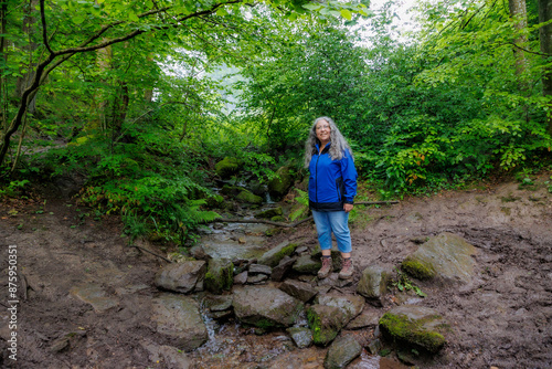 Stream with scant water flowing between rocks, woman hiker standing on stones, mountain in Warche valley, wild vegetation with green foliage on blurred background, summer day in Waimes, Belgium photo