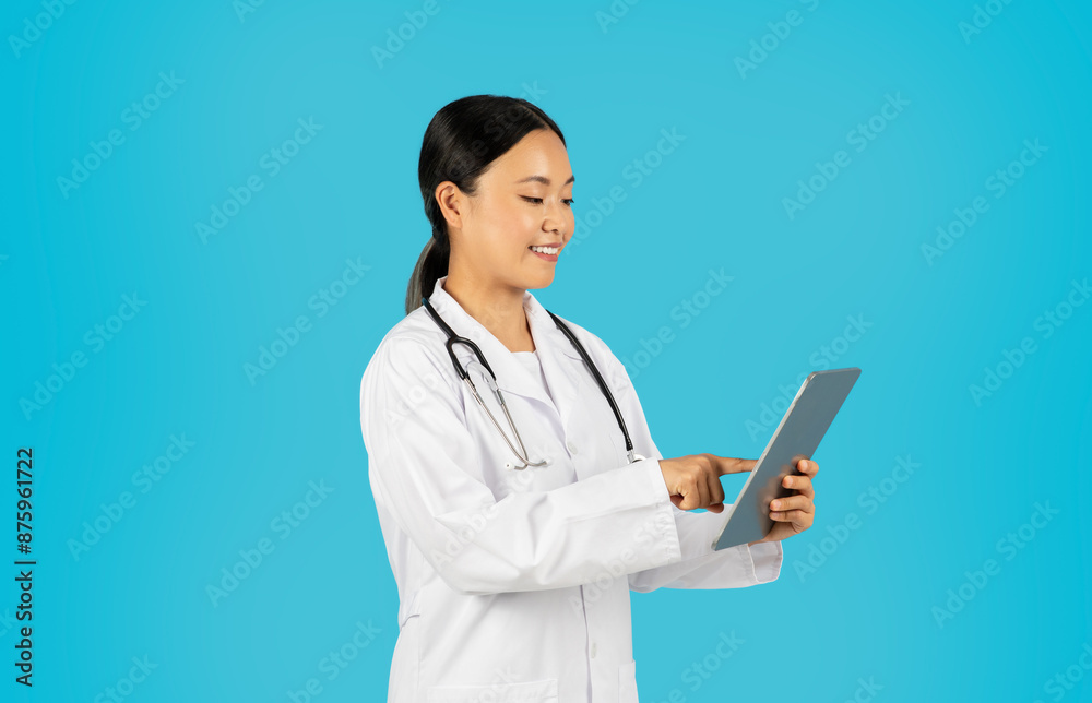 Asian woman doctor, wearing a white lab coat and a stethoscope, stands in front of a blue background, smiling while using a digital tablet. Asian woman doctor, wearing a white lab coat and a stethoscope, stands in front of a blue background, smiling while using a digital tablet.