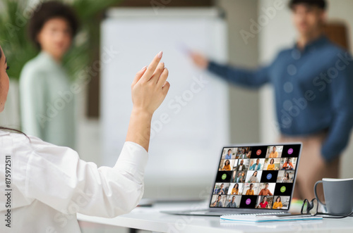 Woman sitting in a meeting, raising her hand to ask a question. The room appears to be a hybrid setting, with the woman in person and other participants appearing on the laptop screen photo