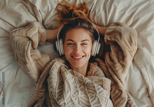 A woman lies in bed with a white blanket, wearing headphones and smiling.