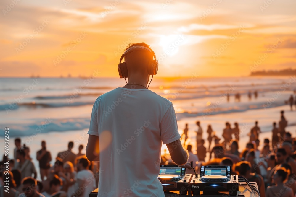A DJ performs in front of a large crowd at a beach party during sunset. The sun is setting over the ocean, and the waves are crashing in the background.