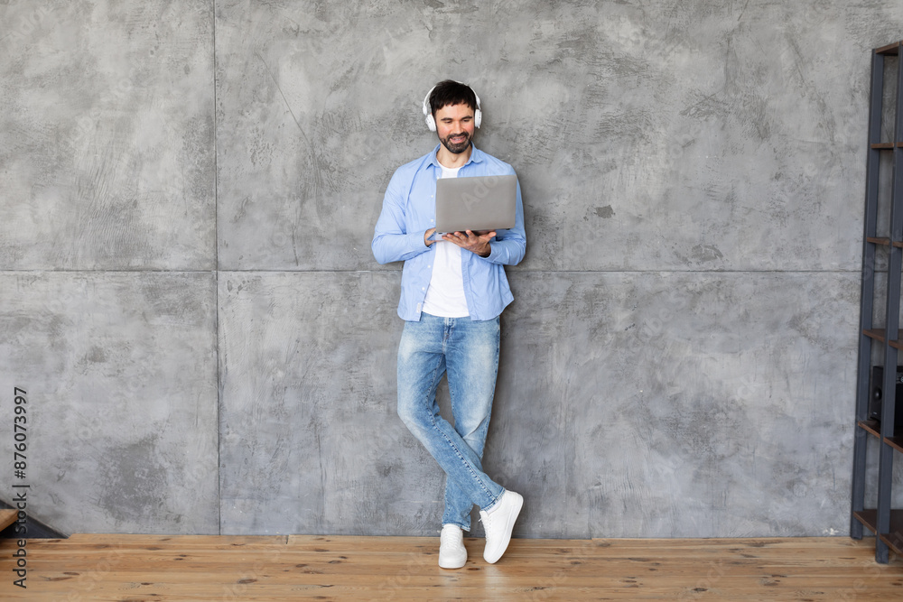 A man stands against a concrete wall, using a laptop computer. He is wearing headphones and a blue button-down shirt, and he is smiling at the camera. He is leaning against the wall A man stands against a concrete wall, using a laptop computer. He is wearing headphones and a blue button-down shirt, and he is smiling at the camera. He is leaning against the wall