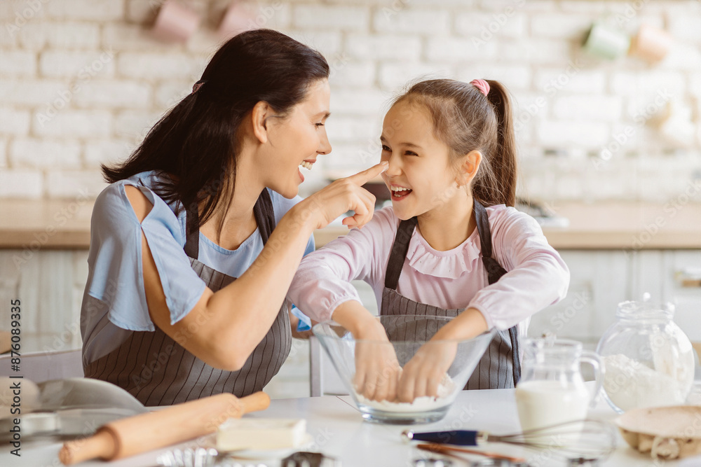 A mother and daughter are baking together in the kitchen. The mother is wearing a blue shirt and a striped apron, while the daughter is wearing a pink shirt and a striped apron A mother and daughter are baking together in the kitchen. The mother is wearing a blue shirt and a striped apron, while the daughter is wearing a pink shirt and a striped apron
