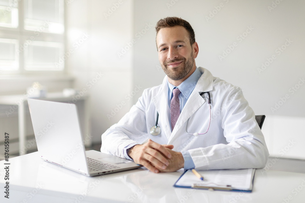 Portrait of professional man general practitioner working on laptop computer in clinic office, preparing to attend appointment, smiling at camera Portrait of professional man general practitioner working on laptop computer in clinic office, preparing to attend appointment, smiling at camera