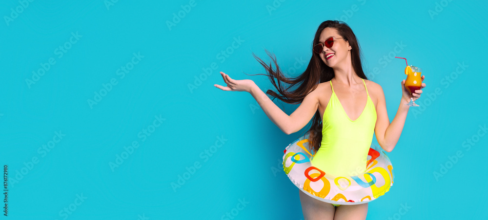 A young woman in a yellow swimsuit holds a colorful cocktail and an inflatable ring while smiling against a bright blue background, copy space