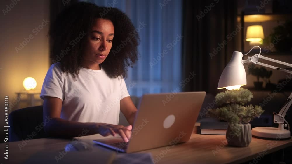 African American woman working on a laptop at a home office, lit by a lamp and surrounded by a plant