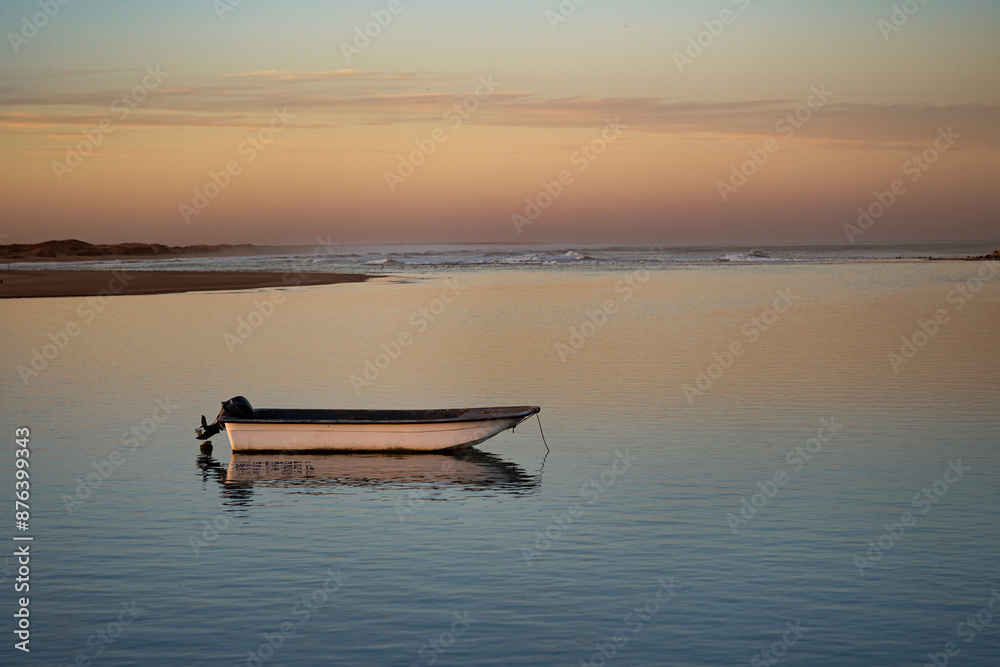 A small boat floats peacefully on calm water at sunset, with a vibrant orange sky reflecting off the water's surface and a distant shoreline silhouetted against the horizon. A small boat floats peacefully on calm water at sunset, with a vibrant orange sky reflecting off the water's surface and a distant shoreline silhouetted against the horizon.