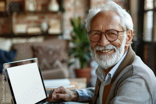 The beautiful senior man, wearing glasses and a well-groomed beard, is smiling while using a laptop with a blank white screen. © Vera