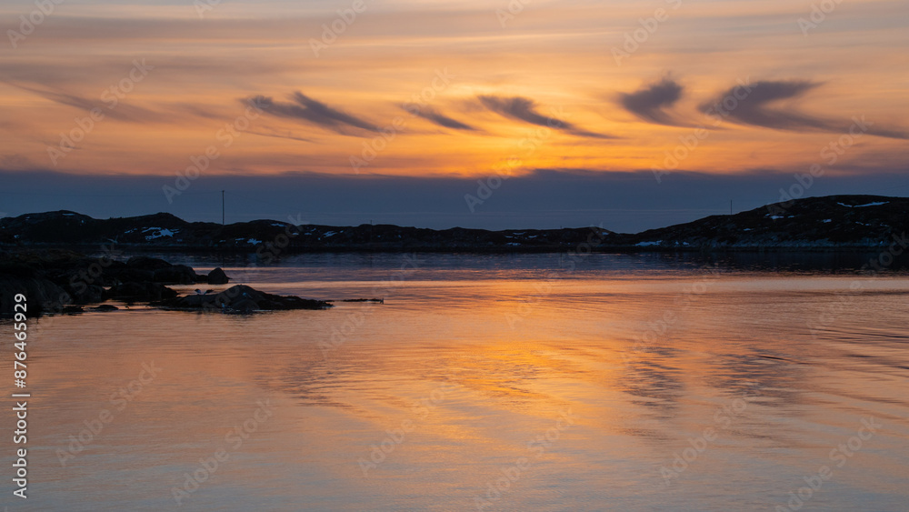 Obraz premium Serene sunset over a calm body of water, with the sky painted in warm hues of orange and yellow. Silhouettes of distant hills and scattered rocks create a tranquil and picturesque landscape