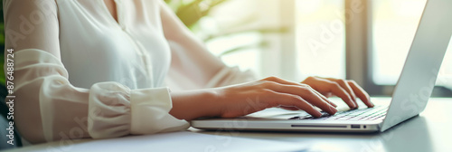 A woman typing on a laptop computer at an office desk, close up view of hands and equipment with a blurred background

