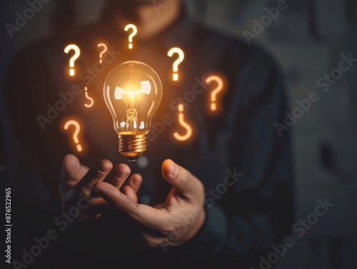 In front of a dark background filled with floating lights and question marks, a hand holds a glowing light bulb.
