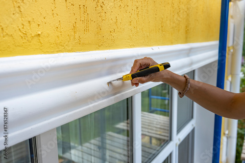 Fotografie A hand is holding a utility knife and is removing excess caulk from a white trim above a window on a sunny day