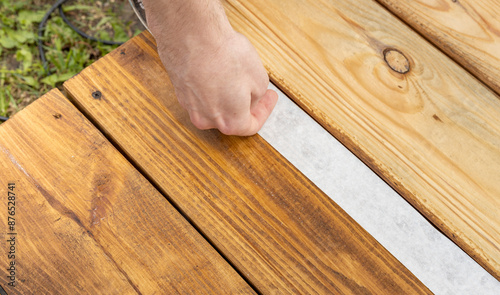 A hand carefully presses down white tape on a freshly stained wooden deck, ensuring a clean line for the next step in the project.