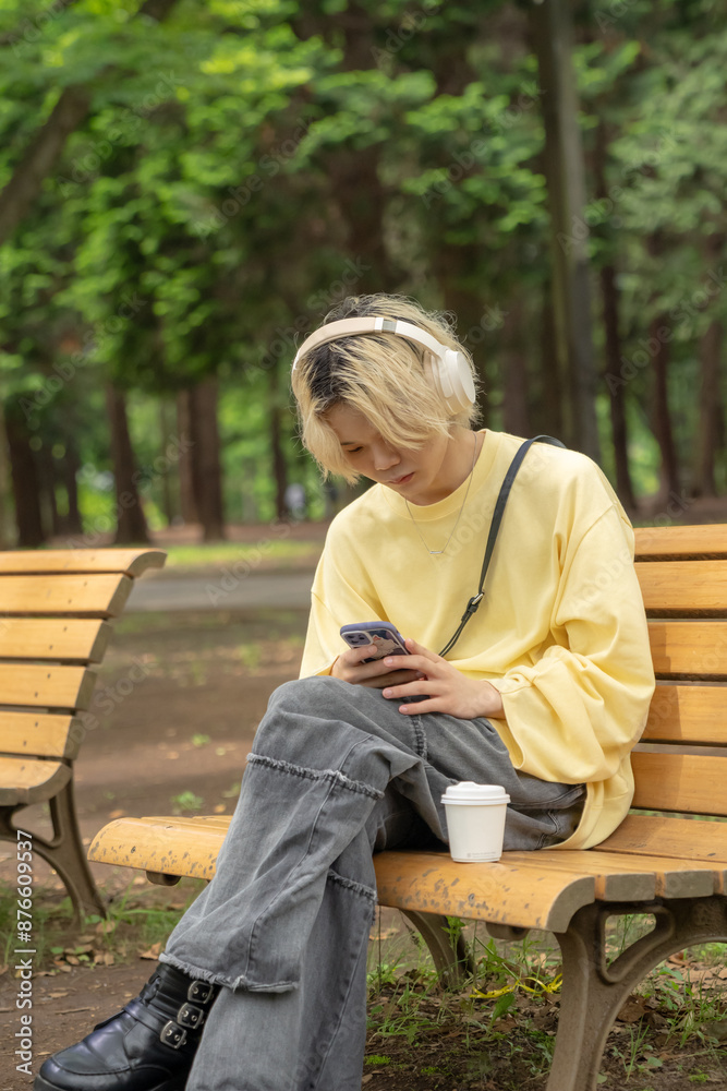 In Yoyogi Park, Shibuya, Tokyo, a Japanese man in his twenties wearing yellow clothes, sitting on a secluded bench, operating a smartphone along with headphones, music, and coffee.