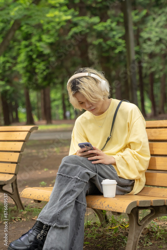 Obraz na płótnie In Yoyogi Park, Shibuya, Tokyo, a Japanese man in his twenties wearing yellow clothes, sitting on a secluded bench, operating a smartphone along with headphones, music, and coffee. w salonie