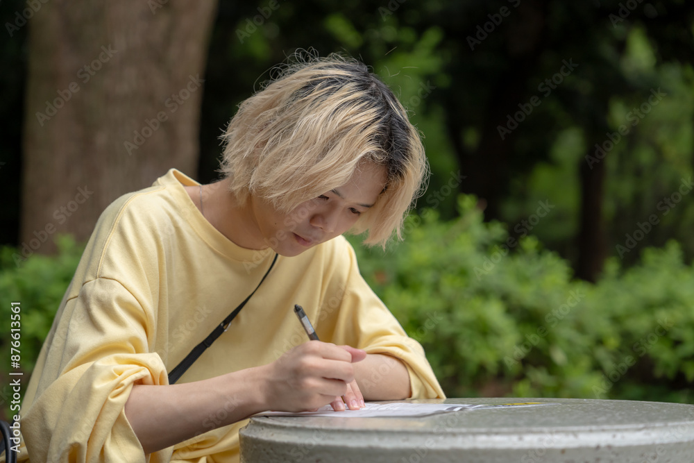 In early summer, a young Japanese man in his early twenties wearing yellow clothes, sitting at a table in Yoyogi Park, Shibuya, Tokyo, writing on paper during the day. In early summer, a young Japanese man in his early twenties wearing yellow clothes, sitting at a table in Yoyogi Park, Shibuya, Tokyo, writing on paper during the day.
