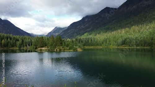 Flying over a Northwest Tranquil Lake Reflecting Dense Forest and Mountain Peaks Under a Cloudy Sky