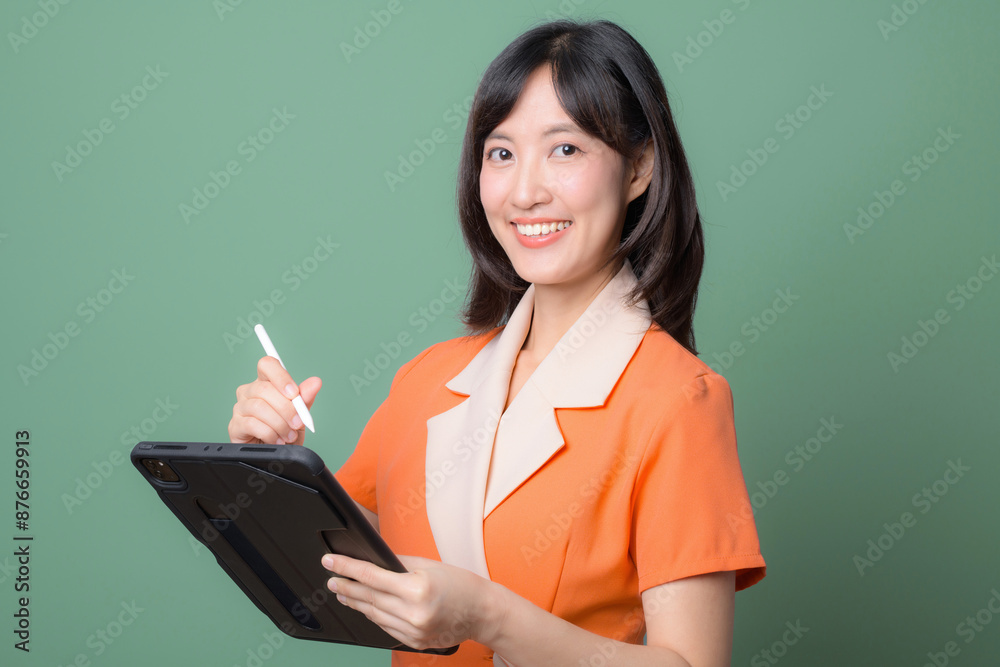 A smiling woman in an orange dress holds a tablet and stylus, posing confidently against a green background. Suitable for business and technology themes. A smiling woman in an orange dress holds a tablet and stylus, posing confidently against a green background. Suitable for business and technology themes.