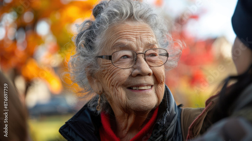 Close-up portrait of a smiling elderly woman with glasses, enjoying a sunny day outdoors with autumn foliage in the background.