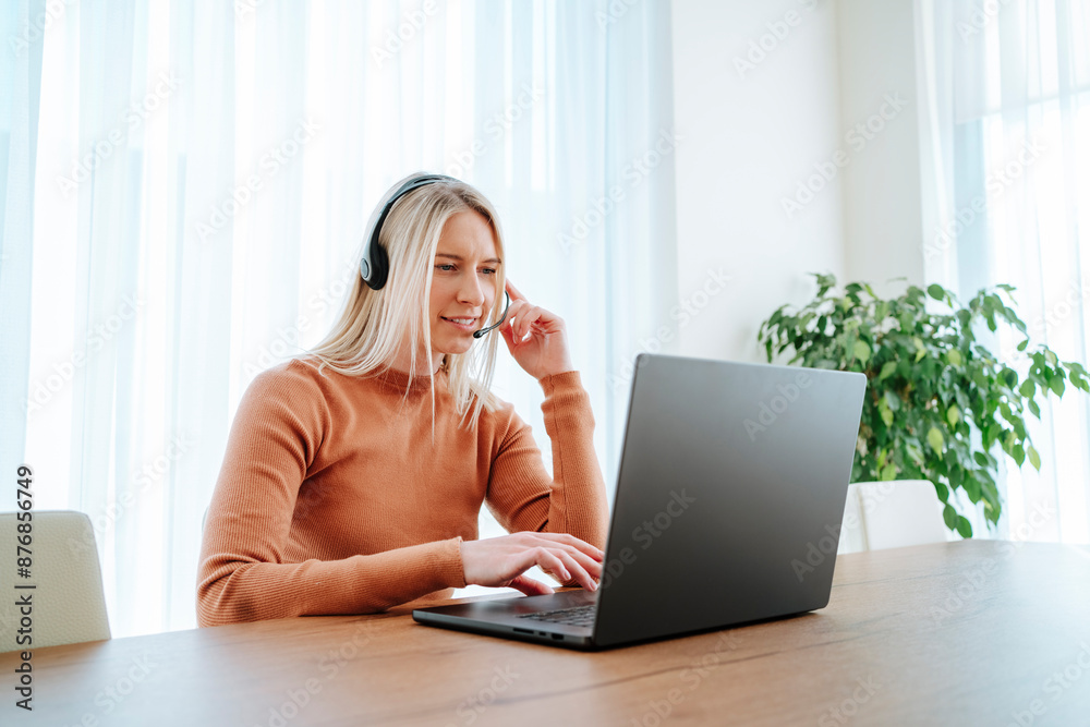 Young blond businesswoman with headset using laptop at table working from home Young blond businesswoman with headset using laptop at table working from home