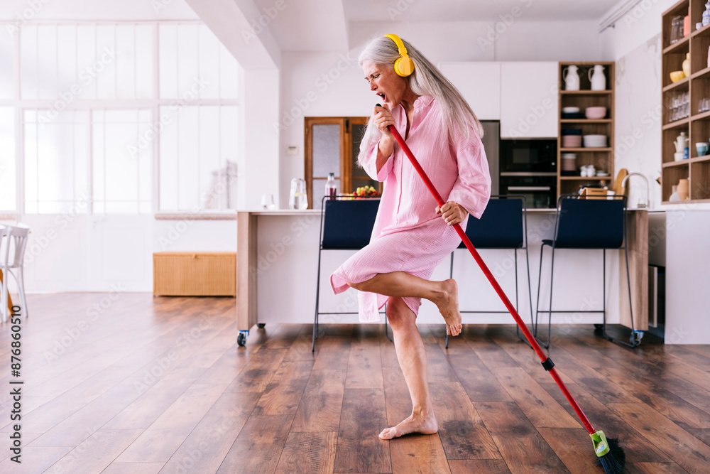 Mature woman with yellow headphones dancing in kitchen with broom Mature woman with yellow headphones dancing in kitchen with broom