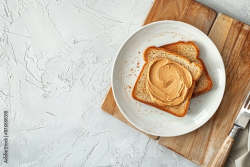 Toast with peanut butter on white ceramic plate with knife on wooden cutting kitchen board, white concrete background, top view flat lay photo