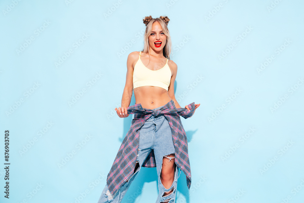 Young beautiful smiling female in trendy summer jeans and top clothes, plaid shirt tied on waist. Carefree woman posing near blue wall in studio. Sexy positive model. Red lips Young beautiful smiling female in trendy summer jeans and top clothes, plaid shirt tied on waist. Carefree woman posing near blue wall in studio. Sexy positive model. Red lips