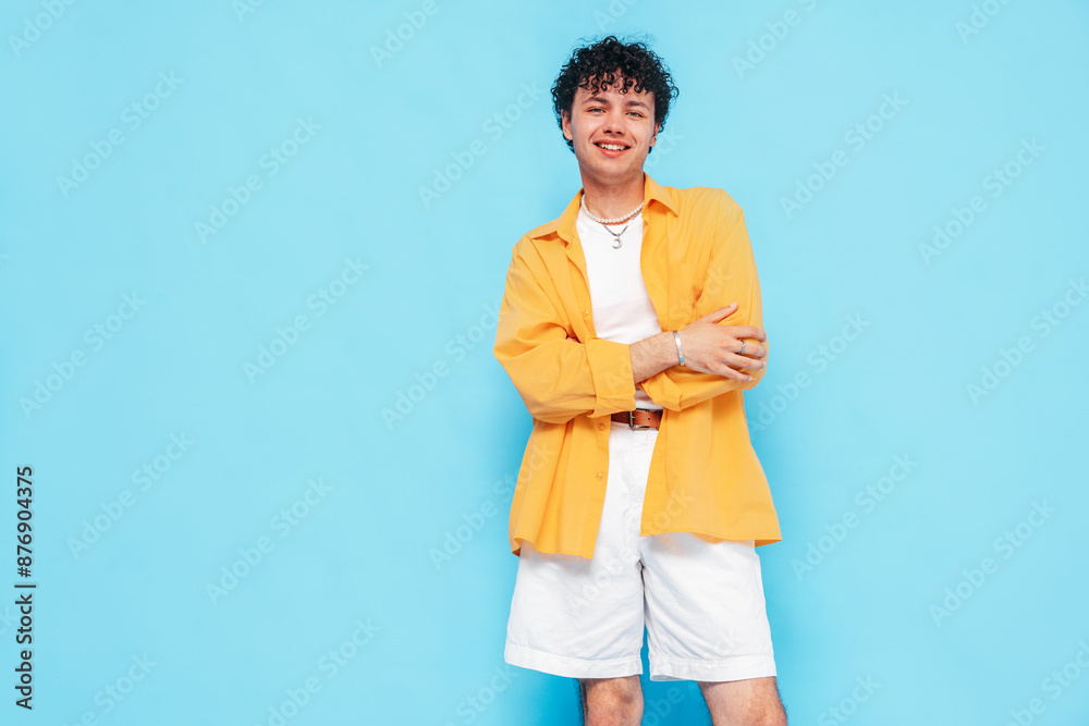 Young attractive man with short curly hair hairstyle. Smiling handsome male in casual stylish yellow shirt and shorts clothes posing in studio, isolated on blue. Cheerful and happy model Young attractive man with short curly hair hairstyle. Smiling handsome male in casual stylish yellow shirt and shorts clothes posing in studio, isolated on blue. Cheerful and happy model