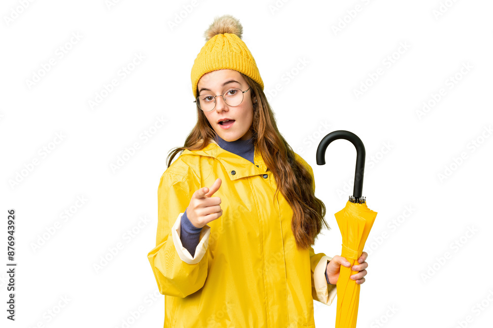 Teenager girl with rainproof coat and umbrella over isolated chroma key background surprised and pointing front Teenager girl with rainproof coat and umbrella over isolated chroma key background surprised and pointing front